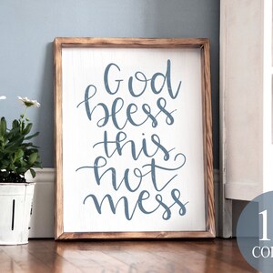 Puede incluir: Un letrero de madera con el texto "God bless this hot mess" en letras azules de escritura cursiva. El letrero está enmarcado en un marco de madera marrón claro y está sentado en un suelo de madera.