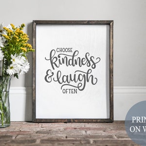 May include: A wooden framed sign with the words "CHOOSE kindness & laugh OFTEN" in black script. The sign is on a white background and has a dark brown frame. A vase of white and yellow flowers sits to the left.