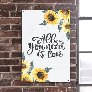 Könnte beinhalten: Ein weißes Metallschild mit einem verwitterten Look, das ein florales Design aus gelben Sonnenblumen mit grünen Blättern zeigt. Das Schild trägt den Text "All you need is love" in schwarzer Kursivschrift.