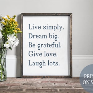 以下が含まれることがあります： 白い背景に青い文字で「Live simply. Dream big. Be grateful. Give love. Laugh lots.」と印刷された、額縁付きの木製看板。看板は木製の表面に置かれ、手前に白と黄色の花が活けられた花瓶がある。