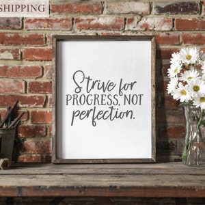 May include: A framed sign with the text "Strive for progress, not perfection." The sign is hanging on a brick wall with a vase of white daisies in front of it.