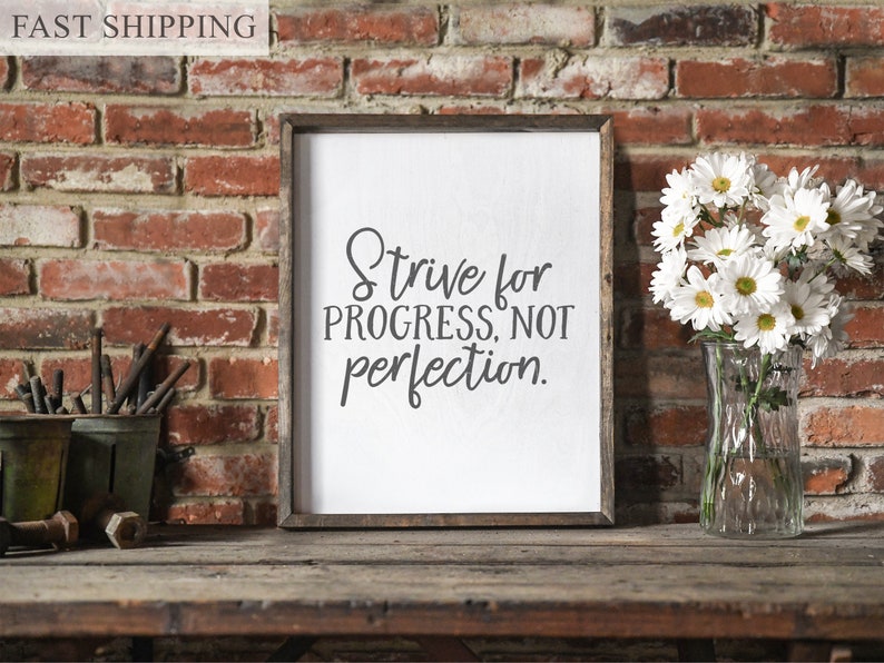May include: A framed sign with the text "Strive for progress, not perfection." The sign is hanging on a brick wall with a vase of white daisies in front of it.