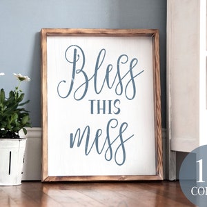 Puede incluir: Un letrero de madera con el texto "Bless this mess" en letras cursivas azules. El letrero está colgado en una pared blanca y tiene un marco de madera rústico.