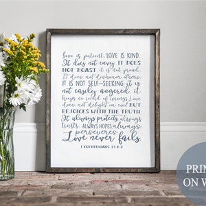 May include: A framed wood sign with a white background and blue text. The text is a quote from the Bible, 1 Corinthians 13:4-7, about love. The sign is printed on wood and has a rustic look. The sign is sitting on a wooden surface with a vase of white flowers in the foreground.