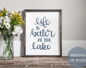 May include: A wooden sign with the text "life is better at the lake" in blue cursive lettering. The sign is framed in a dark wood frame and is sitting on a brick surface. The text "PRINTED ON WOOD" is visible in the lower right corner of the image.