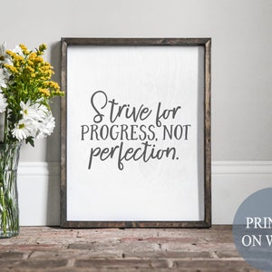 May include: A framed wood sign with the text "Strive for progress, not perfection." printed in black on a white background. The sign is sitting on a brick surface with a vase of white and yellow flowers in the foreground.