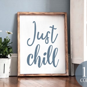 May include: A wooden framed sign with the text "Just chill" in blue lettering on a white background. The sign is leaning against a white dresser.