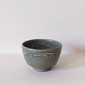 Può includere: Una piccola ciotola in ceramica grigia con un motivo bianco e nero attorno al bordo. La ciotola ha una base arrotondata.