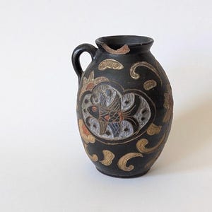 May include: A black ceramic vase with a handle, featuring a detailed design of fish and floral patterns in gold, red, and white. The vase has a rounded body and a slightly flared rim, showcasing an antique aesthetic.
