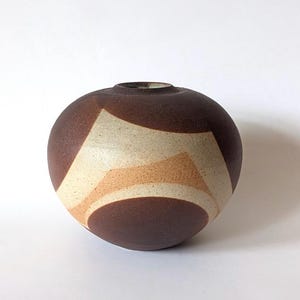 Puede incluir: Un jarrón redondo de cerámica marrón con una pequeña abertura en la parte superior. El jarrón presenta un diseño geométrico de formas crema y beige. La cerámica tiene un acabado mate y se asienta sobre una superficie blanca.