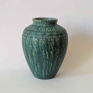 May include: A teal ceramic vase with a textured surface and a geometric pattern around the neck. The vase has a rounded body and a slightly flared opening. The design is reminiscent of vintage pottery.