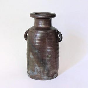 Puede incluir: Un jarrón de cerámica marrón oscuro hecho a mano, con cuerpo cilíndrico y borde ancho y redondeado. El jarrón presenta dos asas pequeñas y curvas y líneas verticales texturizadas. El jarrón mide aproximadamente 25 cm de altura.