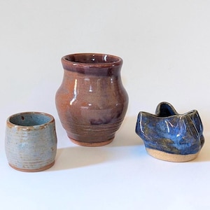 May include: Three handmade ceramic vessels. A small, pale blue cup, a brown vase, and a dark blue, sculptural bowl. The pottery has a rustic, organic aesthetic. The vessels are made of ceramic.