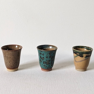 Puede incluir: Tres tazas de cerámica con diferentes patrones de esmalte. La taza de la izquierda tiene un esmalte marrón con motas. La taza del medio tiene un esmalte turquesa con motas negras. La taza de la derecha tiene un esmalte beige con vetas negras y verdes.