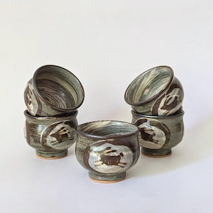 Könnte beinhalten: Ein Set aus sechs Keramikschalen. Die Schalen sind in einem gedämpften Grün gehalten und mit braunen und weißen abstrakten Designs verziert. Das Innere der Schalen weist ein Wirbelmuster auf. Die Schalen sind paarweise gestapelt.