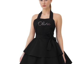 May include: Black apron with a halter neck and a tiered skirt design. The apron features a tie at the waist and the name "Olivia" embroidered in pink script across the chest. The apron is designed for kitchen or craft use.