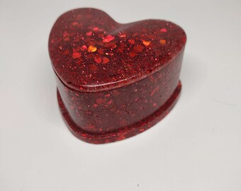 May include: A heart-shaped trinket box with a glossy, glittery red finish. The box is made of a solid material and features a lid that fits snugly on the base. The glitter is a mix of red hues, creating a sparkling effect.