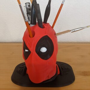 Puede incluir: Porta-bolígrafos con forma de cabeza de Deadpool rojo y negro con una base negra. El organizador tiene varios agujeros para bolígrafos, lápices y pinceles. Hay cinco utensilios de escritura en el organizador.