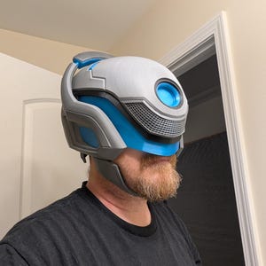 Puede incluir: Un casco blanco y azul con una visera y un gran ojo azul redondo en la parte delantera. El casco está hecho de plástico y tiene una superficie texturizada.