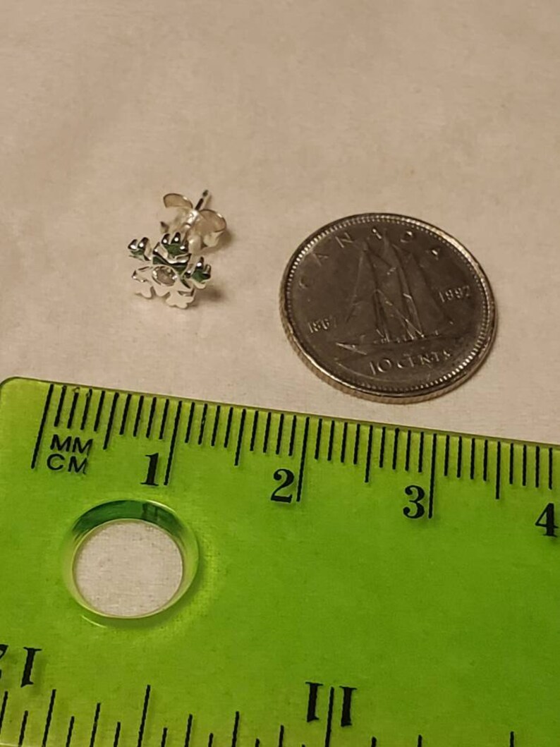 Op de afbeelding: Een zilveren sneeuwvlok oorbel met een heldere strass in het midden. De oorbel is ongeveer 1,25 cm in diameter en wordt getoond naast een Canadese dime ter vergelijking van de grootte.