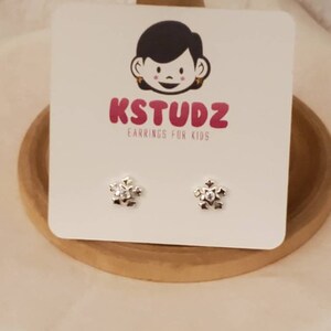 Op de afbeelding: Een paar zilveren sneeuwvlok oorbelletjes voor kinderen. De oorbelletjes zijn op een witte kaart met het logo "KSTUDZ Earrings for Kids".