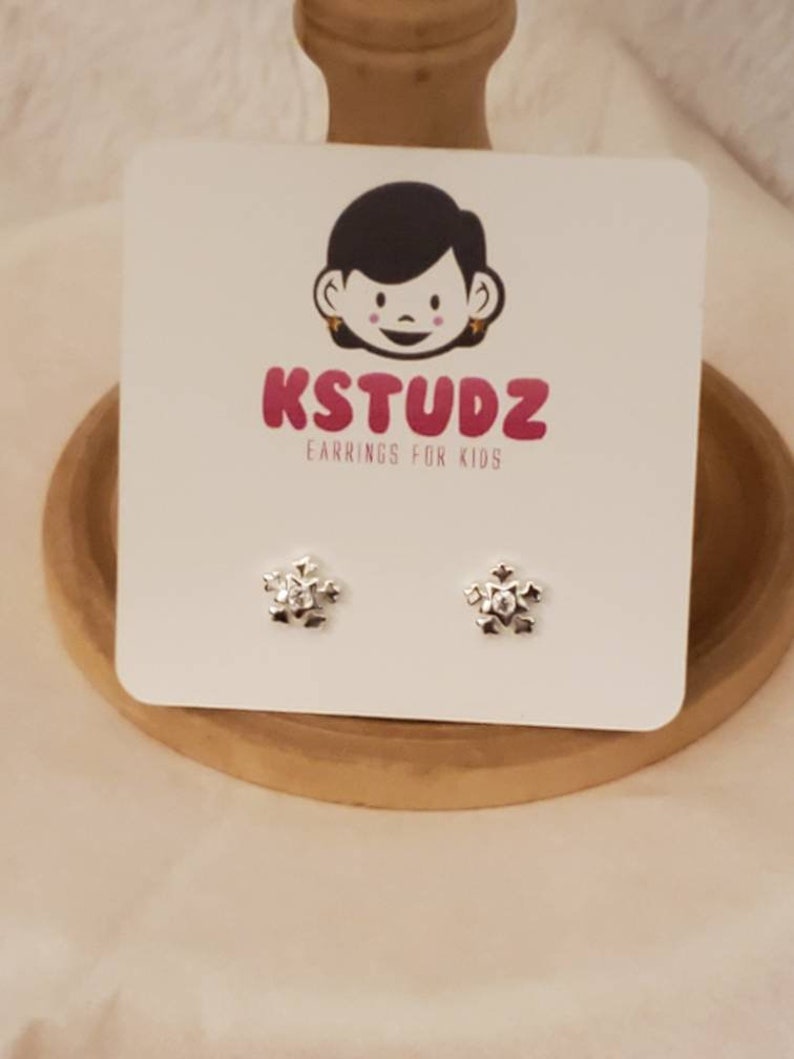 Op de afbeelding: Een paar zilveren sneeuwvlok oorbelletjes voor kinderen. De oorbelletjes zijn op een witte kaart met het logo "KSTUDZ Earrings for Kids".