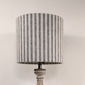 May include: A wooden table lamp with a grey and white striped lampshade. The lamp has a distressed, light grey finish. A glass vase with white roses sits to the left of the lamp. The lamp is approximately 61 cm tall.