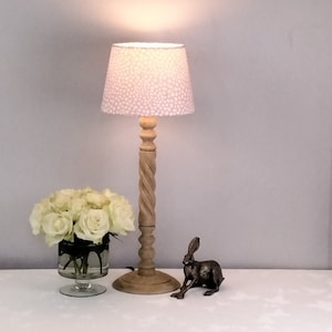 May include: A wooden table lamp with a light pink dotted lampshade. A bouquet of white roses in a glass vase and a bronze rabbit figurine are on the table. The lamp has a twisted wooden base.