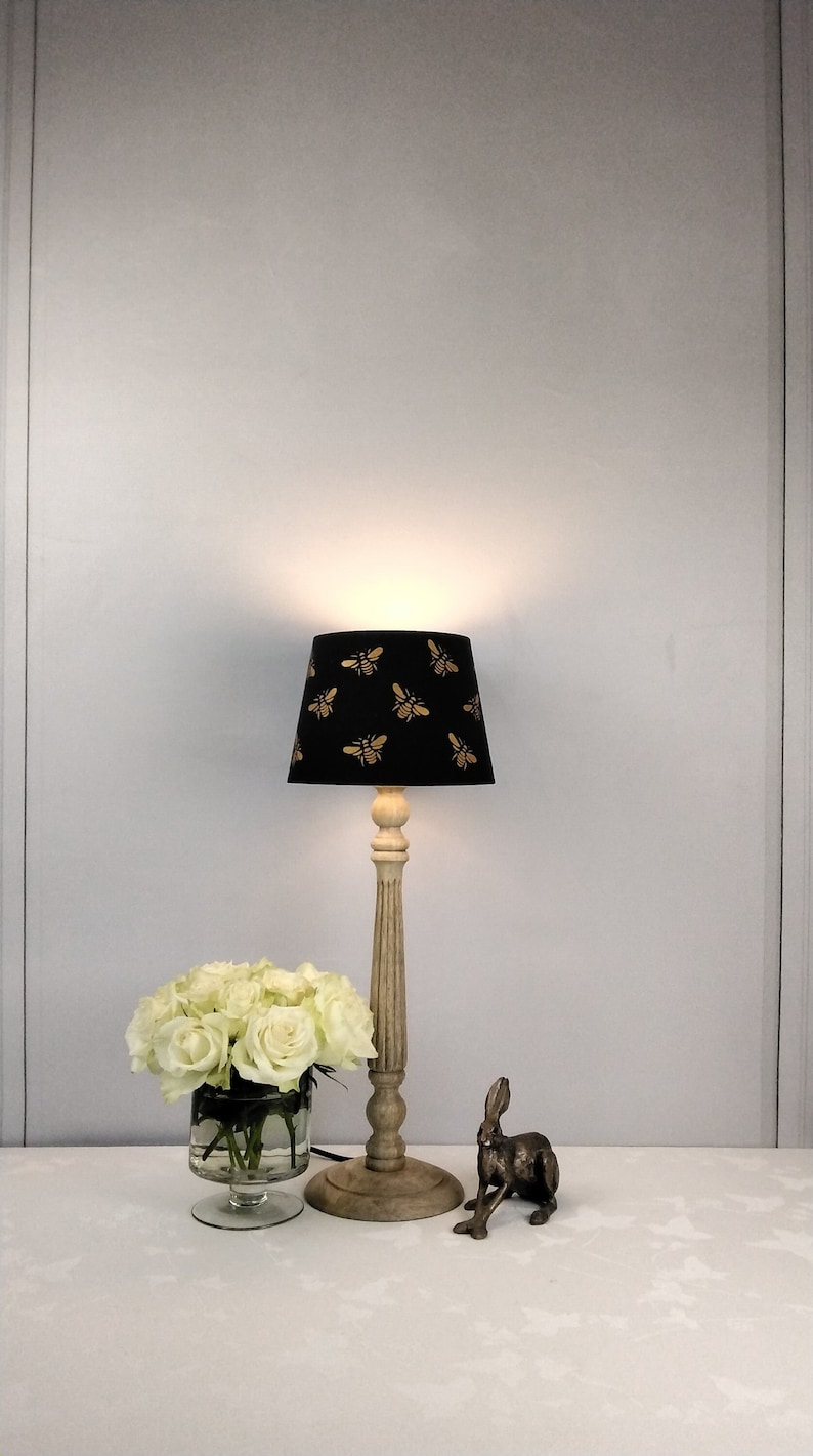 Puede incluir: Una l&aacute;mpara de mesa decorativa con una pantalla negra con un dise&ntilde;o de abejas doradas. La l&aacute;mpara tiene una base de madera clara. Un jarr&oacute;n con rosas blancas y una figura de conejo de bronce tambi&eacute;n est&aacute;n sobre la mesa.