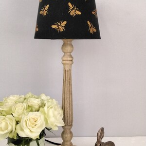 Puede incluir: L&aacute;mpara de mesa decorativa con pantalla negra adornada con motivos de abejas doradas. La l&aacute;mpara tiene una base y un tallo de madera clara. Un ramo de rosas blancas en un jarr&oacute;n y una figura de conejo de bronce tambi&eacute;n est&aacute;n presentes.