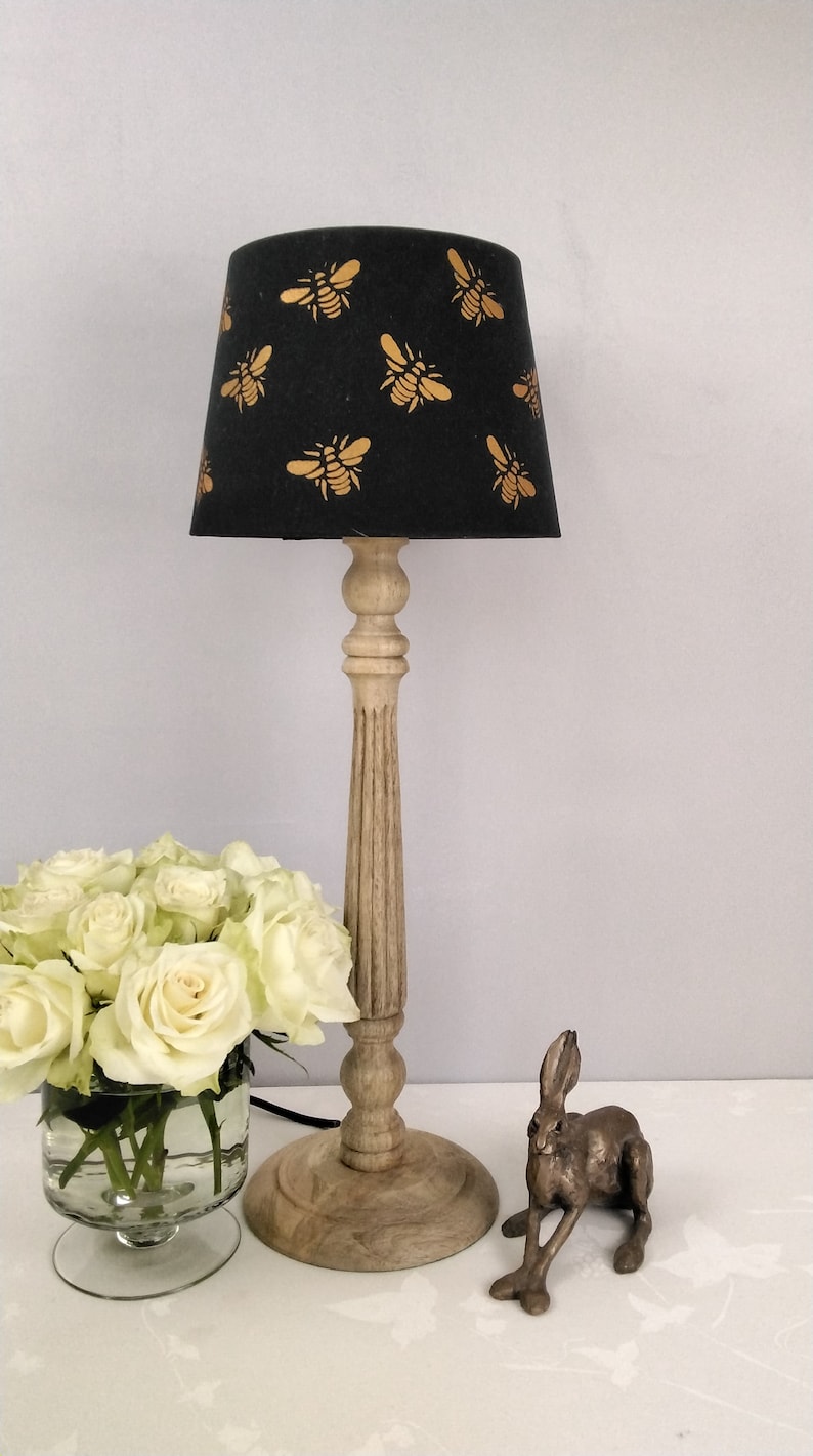 Puede incluir: L&aacute;mpara de mesa decorativa con pantalla negra adornada con motivos de abejas doradas. La l&aacute;mpara tiene una base y un tallo de madera clara. Un ramo de rosas blancas en un jarr&oacute;n y una figura de conejo de bronce tambi&eacute;n est&aacute;n presentes.