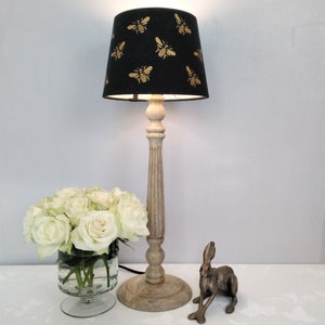 Puede incluir: Una l&aacute;mpara de mesa decorativa con una pantalla negra con un patr&oacute;n de abejas doradas. La l&aacute;mpara tiene una base de madera clara y est&aacute; junto a un ramo de rosas blancas en un jarr&oacute;n de cristal y una figura de conejo de bronce.