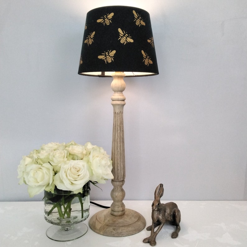 Puede incluir: Una l&aacute;mpara de mesa decorativa con una pantalla negra con un patr&oacute;n de abejas doradas. La l&aacute;mpara tiene una base de madera clara y est&aacute; junto a un ramo de rosas blancas en un jarr&oacute;n de cristal y una figura de conejo de bronce.