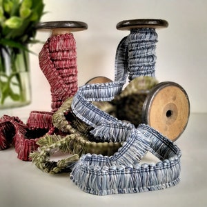 May include: Spools of decorative trim in shades of red, blue, and green. The trim is fringed and wrapped around wooden spools. A vase of green flowers is in the background.