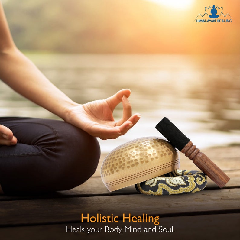 Op de afbeelding: Een persoon mediteert met een gouden zangkom, een zwarte en bruine hamer en een zwart en gouden kussen. De tekst "Holistic Healing Heals your Body, Mind and Soul." staat onder de kom.