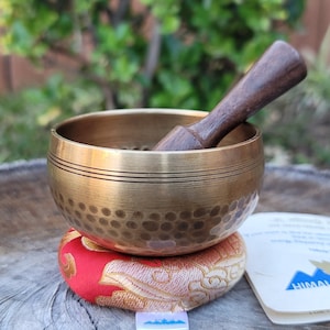 Op de afbeelding: Een zangkom van messing met een houten slager, rustend op een rood en gouden kussen met patronen. De kom heeft een gehammerde textuur en twee dunne banden rond de rand.