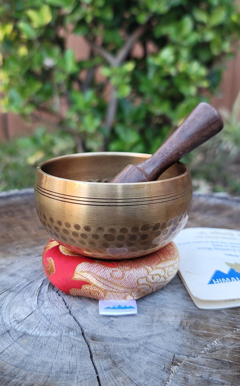Op de afbeelding: Een zangkom van messing met een houten slager, rustend op een rood en gouden kussen met patronen. De kom heeft een gehammerde textuur en twee dunne banden rond de rand.