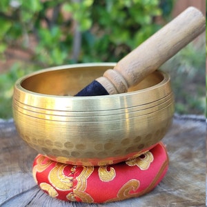Op de afbeelding: Een gouden zangkom met een gehamerde afwerking staat op een rood kussen met gouden borduurwerk. Een houten slager met een zwarte rubberen punt rust op de rand van de kom.