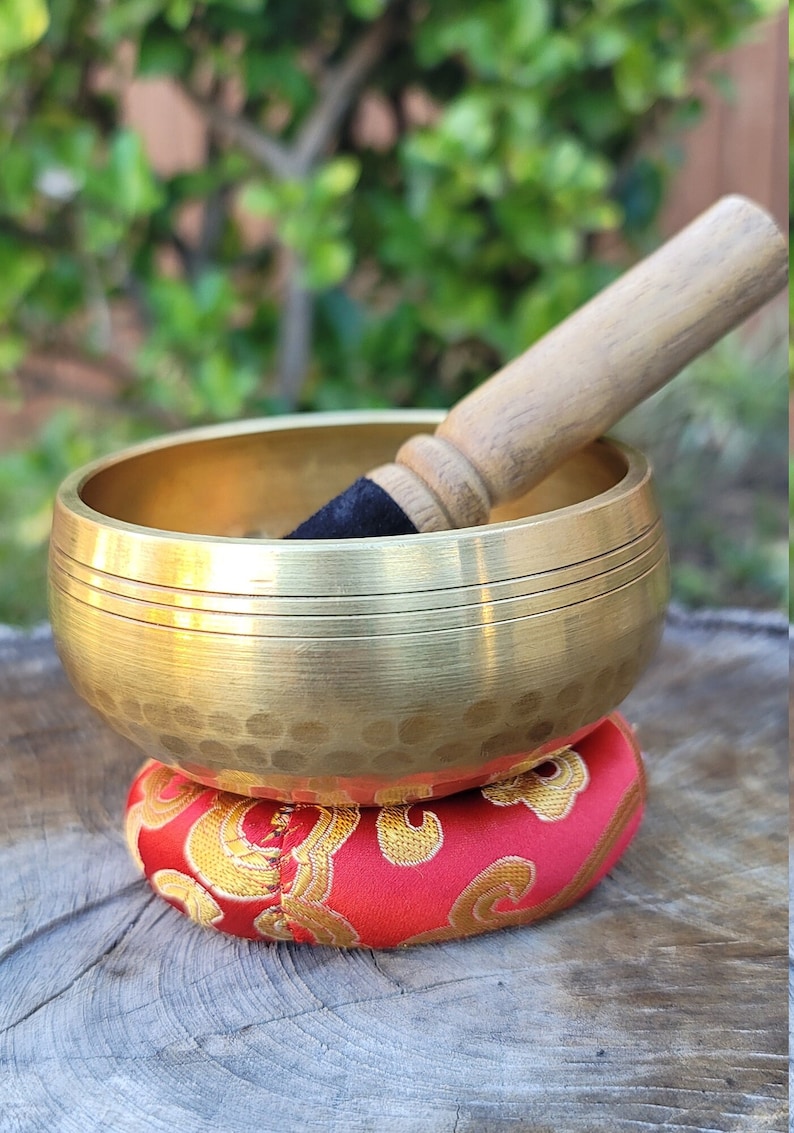 Op de afbeelding: Een gouden zangkom met een gehamerde afwerking staat op een rood kussen met gouden borduurwerk. Een houten slager met een zwarte rubberen punt rust op de rand van de kom.