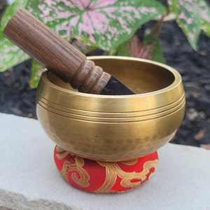 Op de afbeelding: Een gouden zangkom met een gehamerde afwerking en een houten slaagstok. De kom staat op een rood kussen met een gouden patroon.