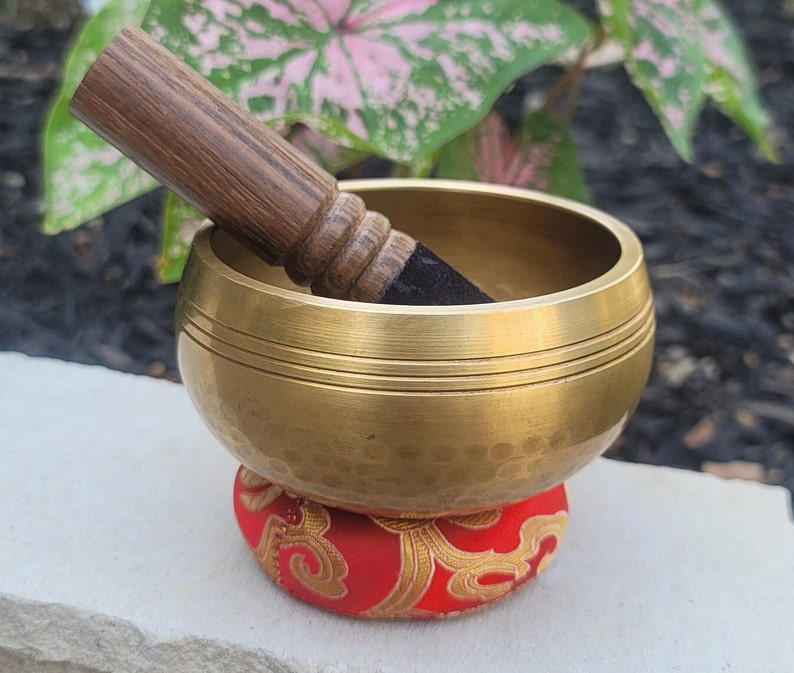 Op de afbeelding: Een gouden zangkom met een gehamerde afwerking en een houten slaagstok. De kom staat op een rood kussen met een gouden patroon.