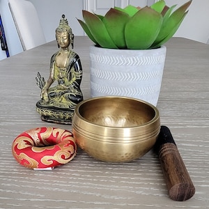 Op de afbeelding: Een gouden zangkom met een houten slaagstok, een rood en gouden meditatiekussen en een klein messing beeldje van een mediterende Boeddha.