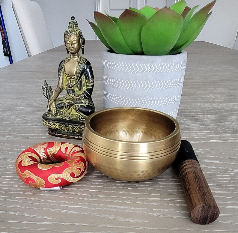 Op de afbeelding: Een gouden zangkom met een houten slaagstok, een rood en gouden meditatiekussen en een klein messing beeldje van een mediterende Boeddha.