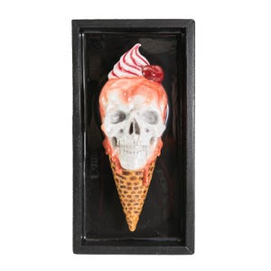 Peut inclure: Sculpture encadrée d'une glace en forme de crâne. Le crâne est blanc avec des détails dégoulinants rouges et oranges, surmonté d'une spirale de crème glacée rose et blanche et d'une cerise. Le cône est marron et le cadre est noir.