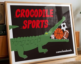 May include: A framed rectangular poster with a black background featuring a green crocodile with its mouth open, holding a basketball, a soccer ball, a baseball, and a football. The text reads CROCODILE SPORTS in red and camelandoasis in white.