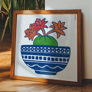 Op de afbeelding: Een vierkante ingelijste print met een gestileerde illustratie van een groene cactus met oranje en rode bloemen in een blauwe kom met witte geometrische patronen, waaronder driehoeken, vierkanten en golvende lijnen.