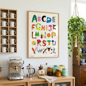 May include: A framed educational alphabet poster featuring colorful illustrations for each letter from A to Z, displayed on a white wall above a wooden counter with a silver espresso machine, a glass French press, and various glass jars.