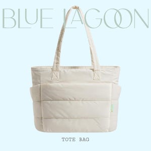 Könnte beinhalten: Cremefarbene Tragetasche mit Steppdesign und zwei Griffen. Die Tasche hat eine rechteckige Form mit einer großen Tasche auf der Vorderseite. Die Worte "BLUE LAGOON" sind oben und "TOTE BAG" unten aufgedruckt.
