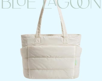 Könnte beinhalten: Cremefarbene Tragetasche mit Steppdesign und zwei Griffen. Die Tasche hat eine rechteckige Form mit einer großen Tasche auf der Vorderseite. Die Worte "BLUE LAGOON" sind oben und "TOTE BAG" unten aufgedruckt.