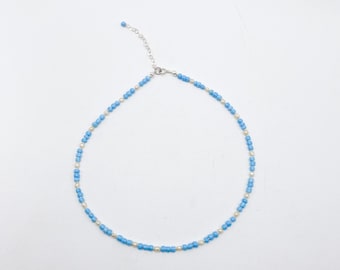May include: A delicate beaded necklace featuring alternating light blue and white beads. The necklace has a silver-toned chain and clasp. The beads are small and evenly spaced, creating a simple yet elegant design. The necklace is displayed on a white background.